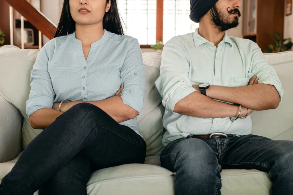 Crop faceless young ethnic couple in casual wear sitting on couch with arms crossed. Observing the habits of a serial cheater can help identify serial cheating or show red flags of serial cheaters.