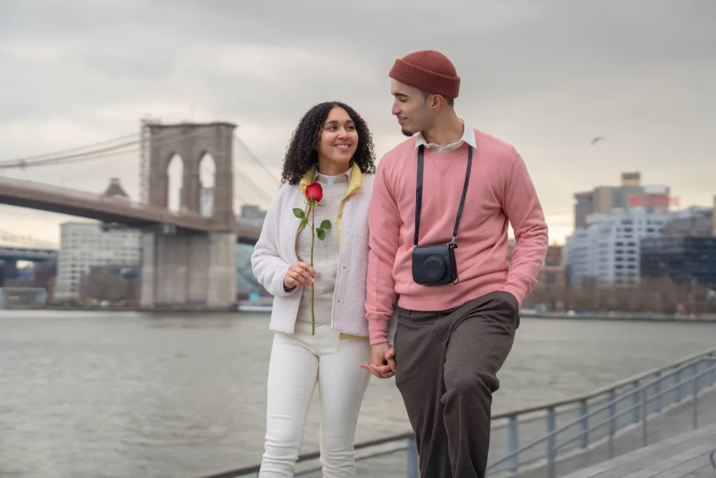 Positive Hispanic girlfriend with rose and boyfriend with photo camera holding hands and looking at each other while strolling on waterfront near sea. Is holding hands part of a farewell kiss, goodbye kisses, or farewell kisses in relationships.