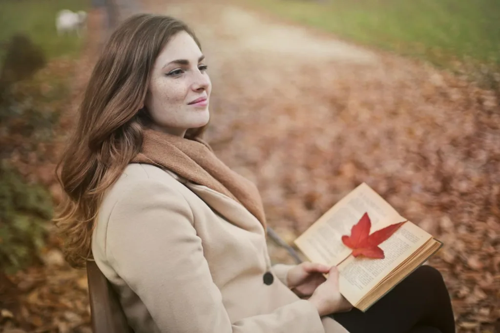 Side view of peaceful young female in warm coat and scarf sitting on bench with open book in hands and looking away pensively while resting in autumn park. Releasing someone uninterested, cutting off communication.