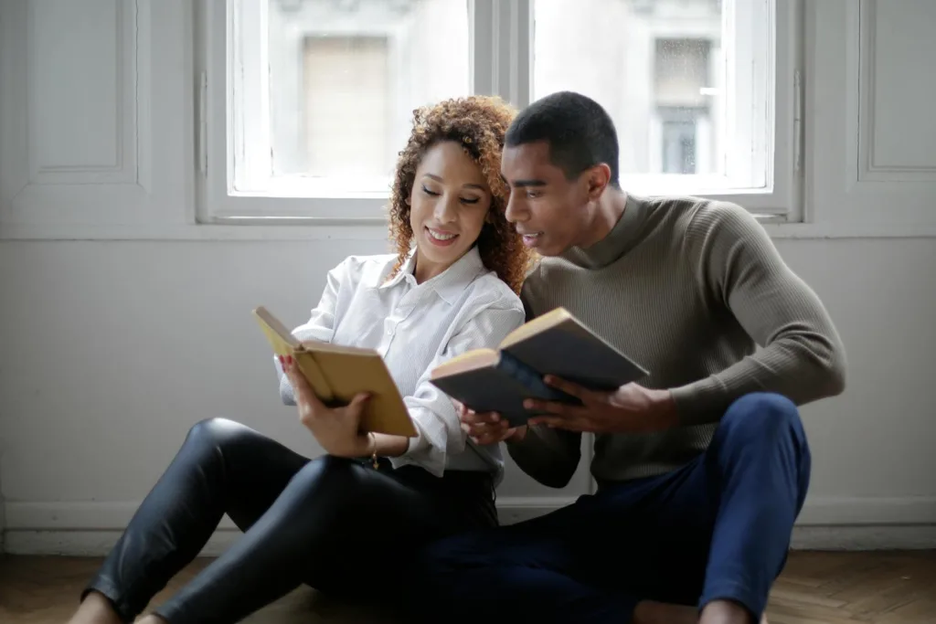 Positive young ethnic couple reading books while sitting on floor near window. Rekindling romance, open communication. help your boyfriend, relationship with God
