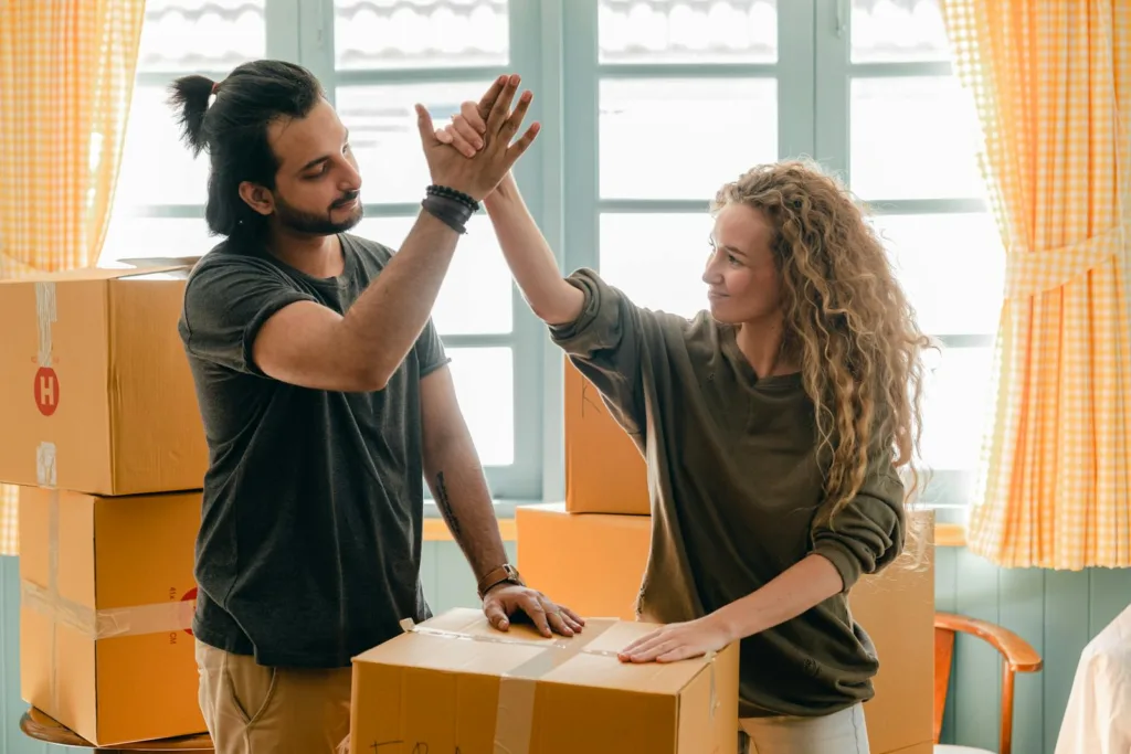 Happy woman in casual wear standing near heap of cardboard boxes and giving high five to ethnic boyfriend with ponytail showing agreement while looking at each other. Rekindling romance, busy couples