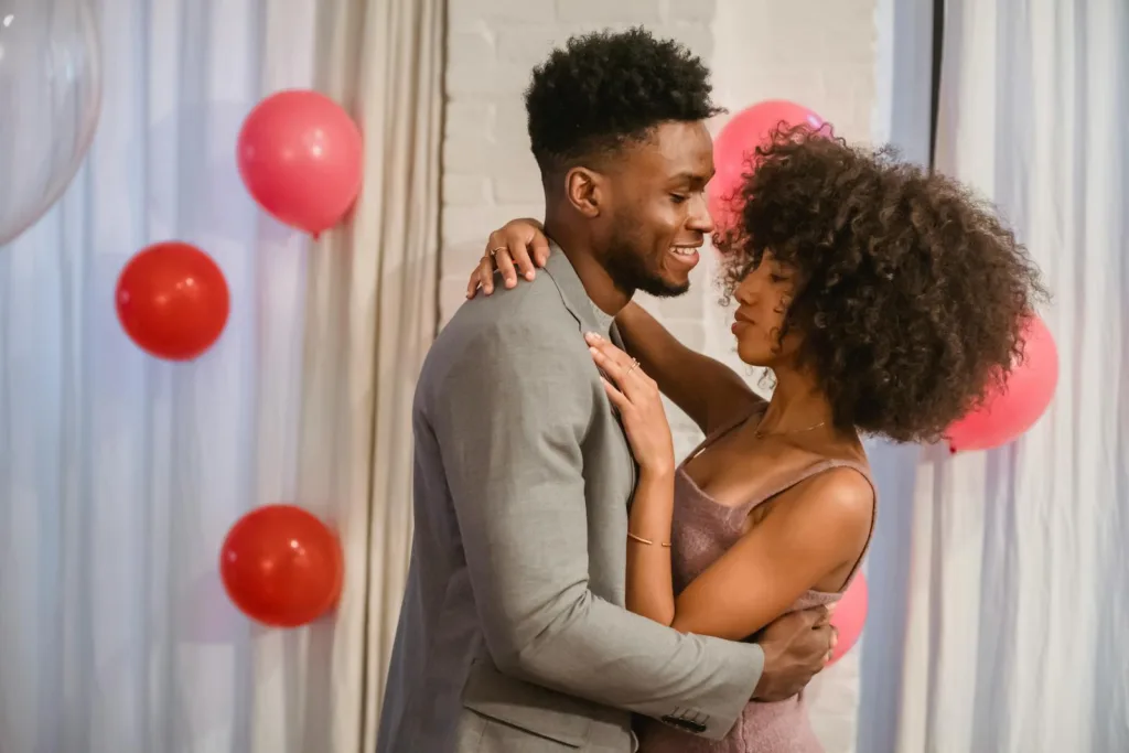 Side view of young black couple in elegant clothes standing in bright room and cuddling while dancing together with closed eyes near pink balloons on wall and curtains. Long-term couples, spark romance