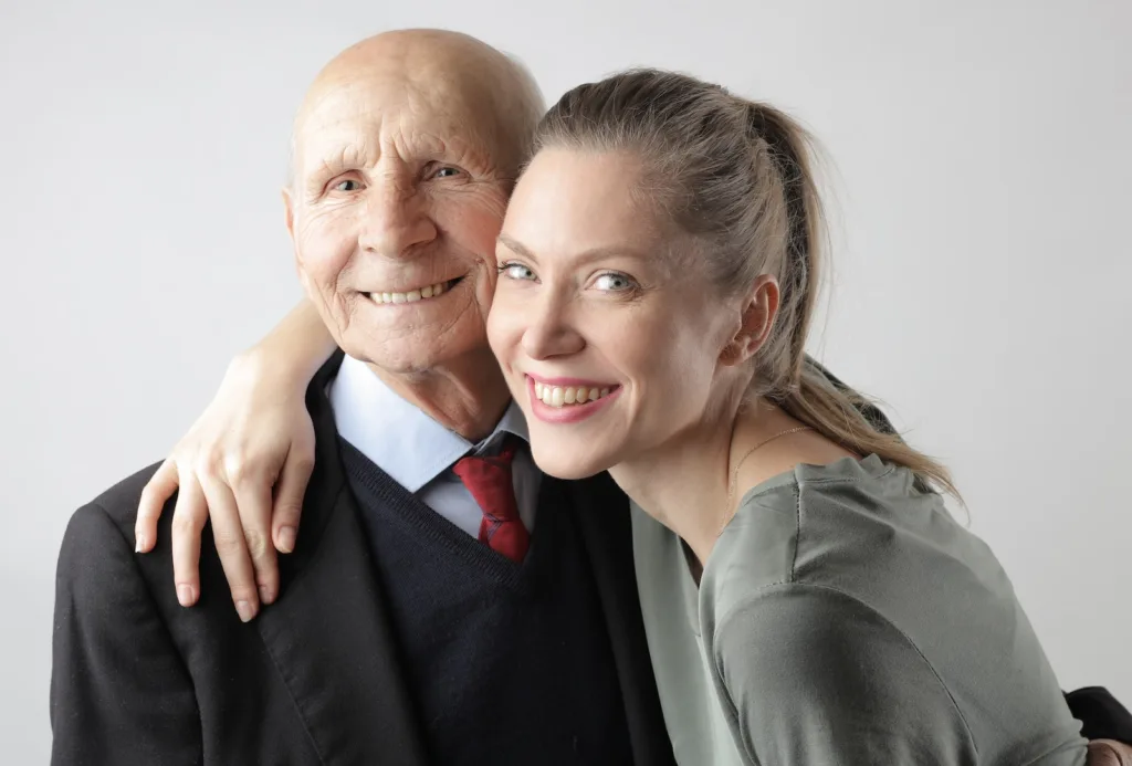 Cheerful elderly man and adult woman cuddling on white background and looking at camera