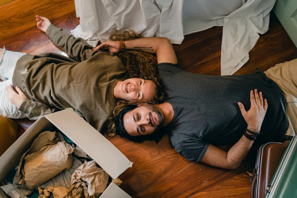 From above of young smiling diverse couple in casual clothing relaxing on floor near carton boxes while unpacking belongings in new contemporary apartment