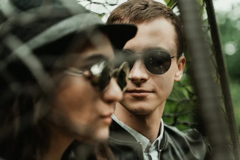 Through mesh fence attractive young man looking at charming woman in stylish hat and sunglasses while enjoying each other