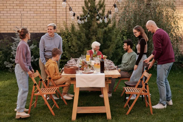 Happy family members talking and sitting down to eat tasty food at big wooden table in backyard in daytime