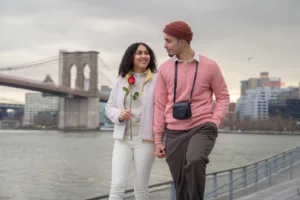 Positive Hispanic girlfriend with rose and boyfriend with photo camera holding hands and looking at each other while strolling on waterfront near sea