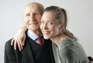 Cheerful elderly man and adult woman cuddling on white background and looking at camera. Attracted to older men, romantic attraction.