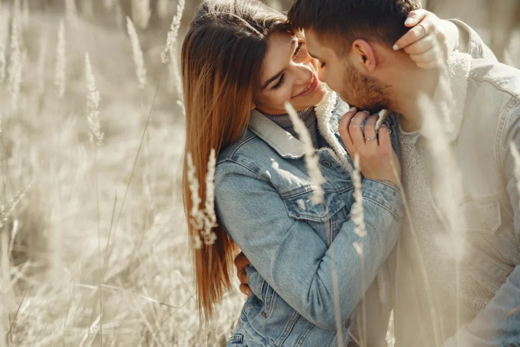 Romantic young couple in warm denim jackets embracing while sitting on dry grass on sunny field in countryside and looking at each other. Overcoming Jealousy, Insecurity in a relationship