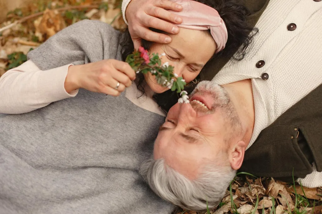 From above of cheerful senior wife wearing wide scarf and headband with flower bouquet in hand and happy elderly gray haired husband in warm clothes lying on ground with fallen leaves in park with closed eyes. Long-term couples, Spark romance