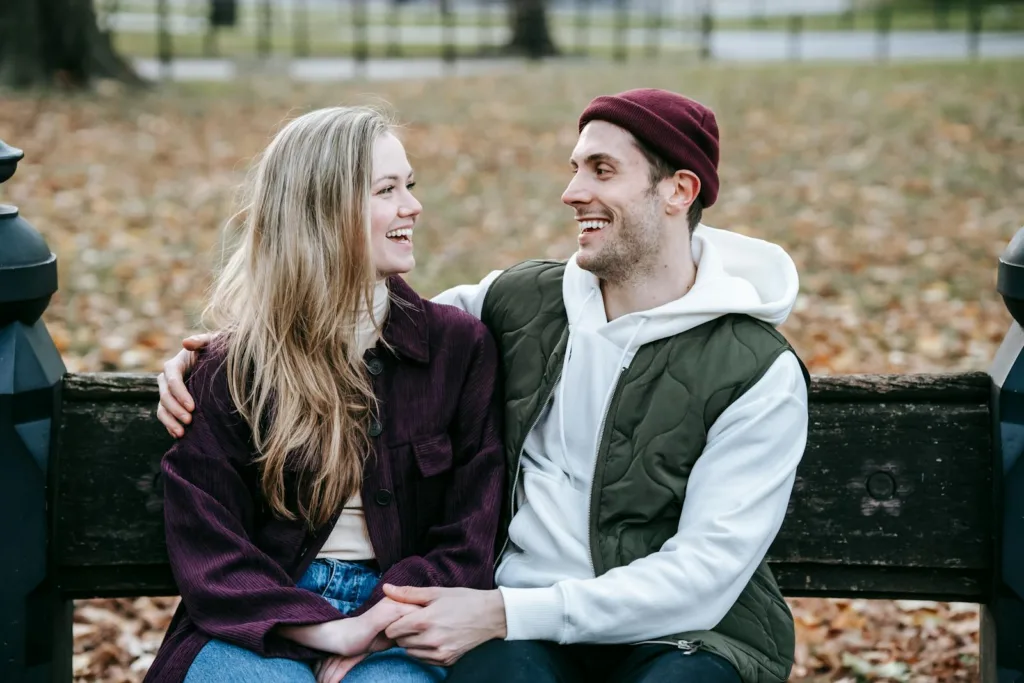 Young happy couple smiling and looking at each other on bench in park with dry autumn leaves. Building trust, romantic relationship. God ordains, natural relationship, balance your relationship, open communication