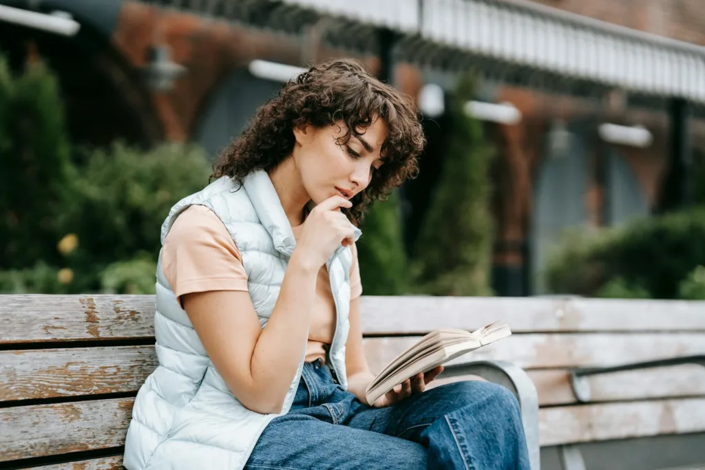Adult thoughtful female touching lips while reading textbook and sitting with crossed legs on wooden bench in town. Cutting off communication, Releasing someone uninterested.