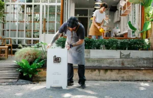 Couple of coworkers in aprons and casual clothes watering plants and putting signboard near cafeteria in daytime. Gratitude to your partner, gestures of love.