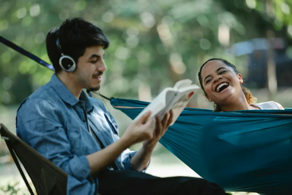 Cheerful young multiracial couple having conversation while resting in forest with book and hammock. Likes and dislikes, deeper emotional connection